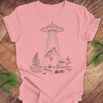 A light pink short-sleeve t-shirt with a black line art graphic of a flying bird under a UFO, displayed flat on a rustic wood background with pine branches.