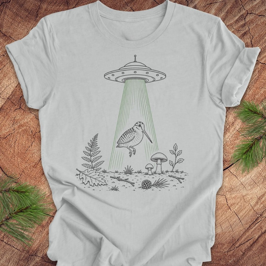 A light gray short-sleeve t-shirt with a black line art graphic of a bird under a UFO with green rays, laid flat on a rustic wood background with pine branches.