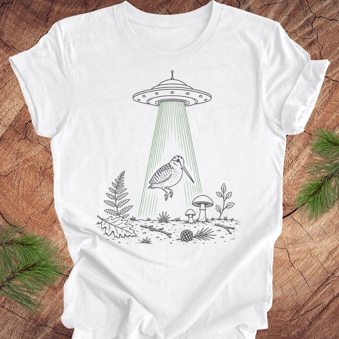 A white short-sleeve crewneck t-shirt laid flat on a rustic wood background, featuring a black line art graphic of a bird under a UFO with green beam rays.