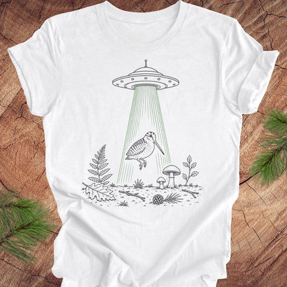 A white short-sleeve crewneck t-shirt laid flat on a rustic wood background, featuring a black line art graphic of a bird under a UFO with green beam rays.