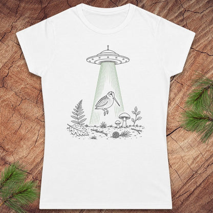 A white short-sleeve crewneck t-shirt featuring a black line art graphic of a UFO with a green beam of light shining down onto a bird, ferns, and mushrooms, laid flat on a rustic wood background with pine branches in the corners.