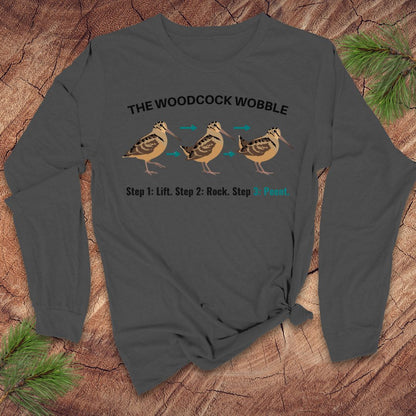 Gray long-sleeve shirt with woodcock design and text on a wooden surface with pine branches.