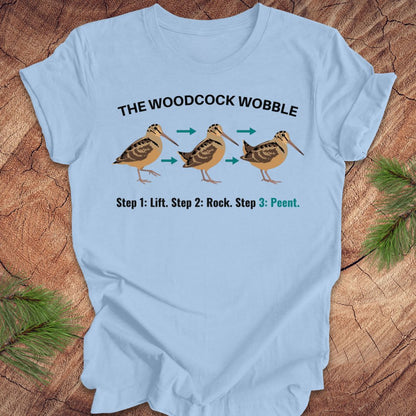 Light blue t-shirt with woodcock wobble graphic and text on a wooden surface.