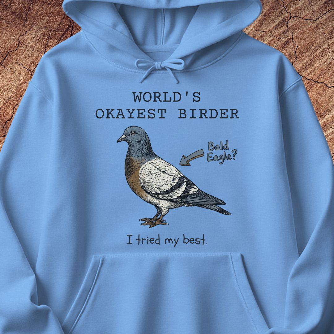 A light blue hoodie laid flat on a rustic wood background, featuring a printed graphic of a pigeon with the text 'WORLD'S OKAYEST BIRDER', 'Bald Eagle?', and 'I tried my best.'.