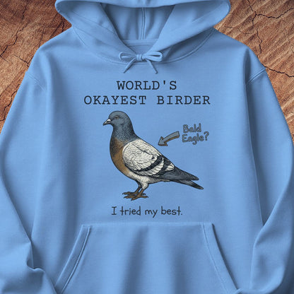 A light blue hoodie laid flat on a rustic wood background, featuring a printed graphic of a pigeon with the text 'WORLD'S OKAYEST BIRDER', 'Bald Eagle?', and 'I tried my best.'.