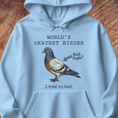 A light blue hoodie laid flat on a textured brown wood background, featuring a printed illustration of a pigeon with the text 'WORLD'S OKAYEST BIRDER' above and 'I tried my best.' below, plus a small arrow reading 'Bald Eagle?'.