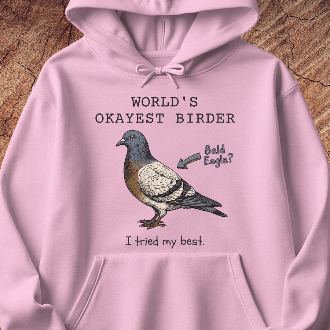 A light pink pullover hoodie laid flat on a textured wood background, featuring a printed graphic of a pigeon with the text 'WORLD'S OKAYEST BIRDER' above it and 'I tried my best.' below it, plus an arrow pointing to the bird that says 'Bald Eagle?'.