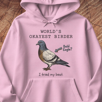 A light pink pullover hoodie laid flat on a textured wood background, featuring a printed graphic of a pigeon with the text 'WORLD'S OKAYEST BIRDER' above it and 'I tried my best.' below it, plus an arrow pointing to the bird that says 'Bald Eagle?'.