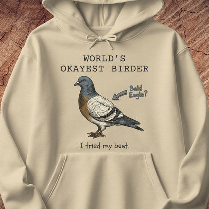 A beige pullover hoodie with a graphic print of a pigeon and humorous text reading 'WORLD'S OKAYEST BIRDER' and 'I tried my best.' The hoodie is displayed flat against a textured brown background.
