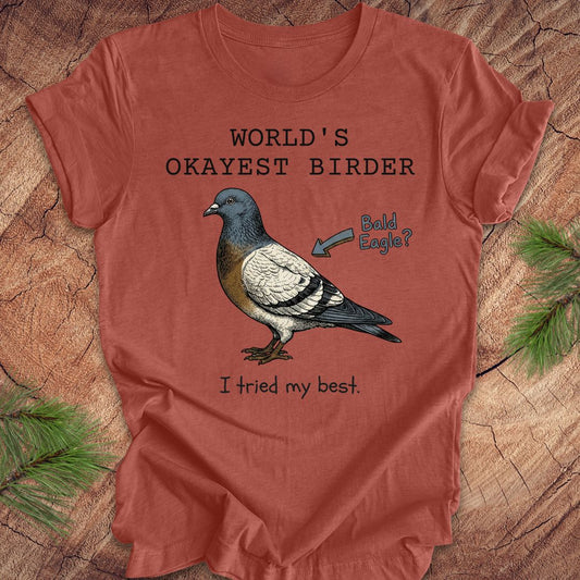 Clay colored t-shirt with a pigeon illustration and text, 'world's okayest birder' on a wooden surface