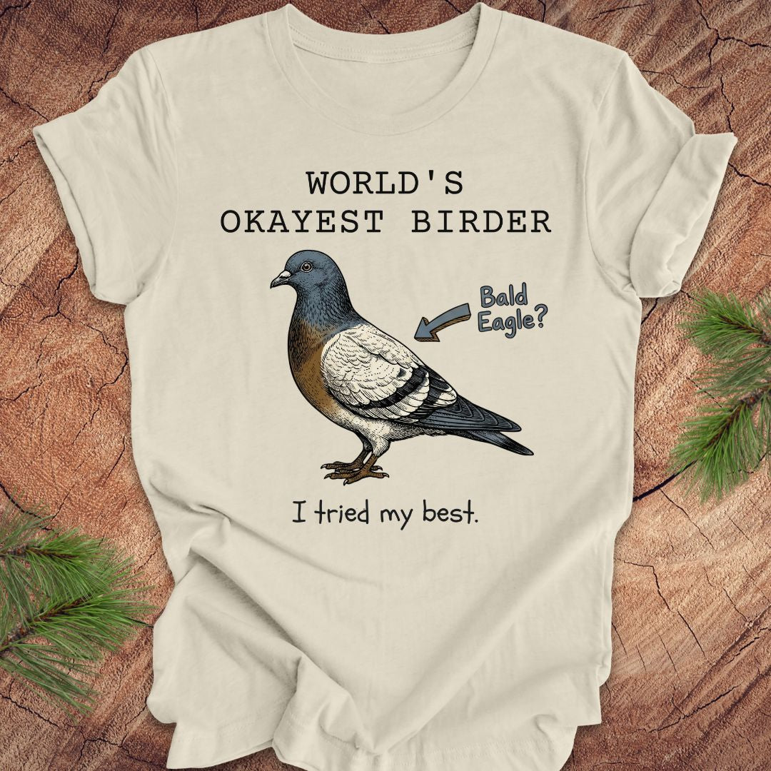 Beige t-shirt with a pigeon illustration and text, 'world's okayest birder' on a wooden surface