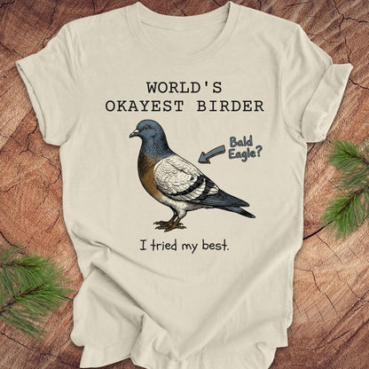 Beige t-shirt with a pigeon illustration and text, 'world's okayest birder' on a wooden surface