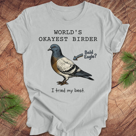 Silver  t-shirt with a pigeon illustration and text, 'world's okayest birder' on a wooden surface