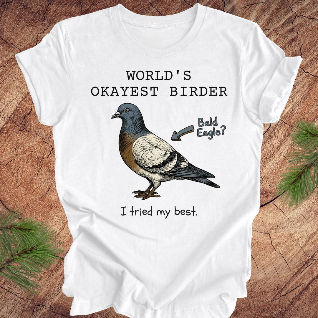 White t-shirt with a pigeon illustration and text, 'world's okayest birder' on a wooden surface
