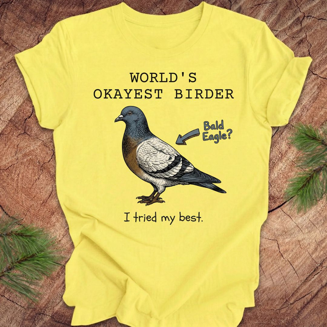 Yellow t-shirt with a pigeon illustration and text, 'world's okayest birder' on a wooden surface