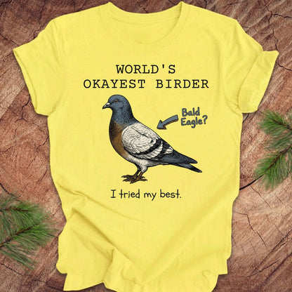 Yellow t-shirt with a pigeon illustration and text, 'world's okayest birder' on a wooden surface