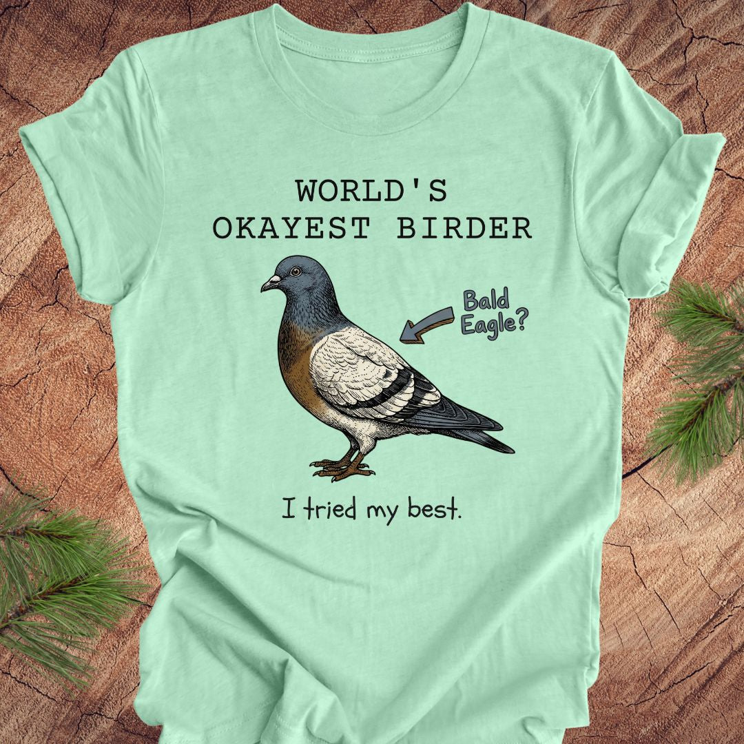 Mint green t-shirt with a pigeon illustration and text, 'world's okayest birder' on a wooden surface
