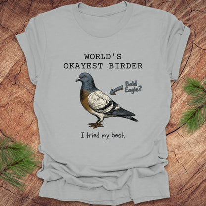 Silver t-shirt with a pigeon graphic and text, "world's okayest birder' on a wooden surface