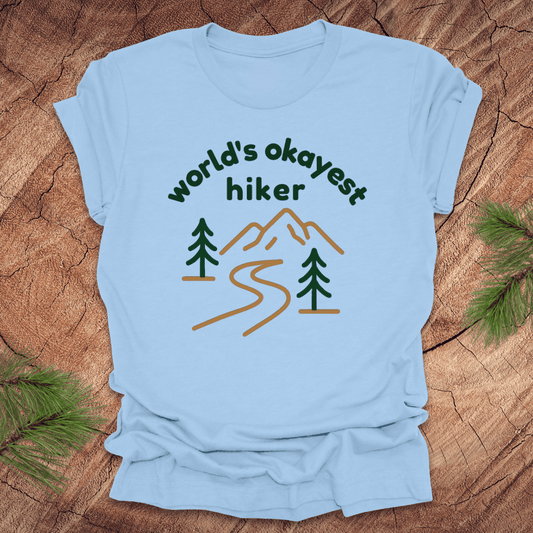 Light blue t-shirt with 'world's okayest hiker' graphic on a wooden surface with pine branches.