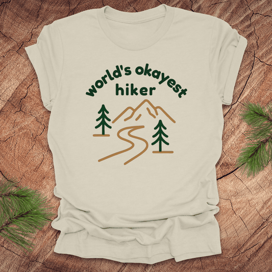 Beige t-shirt with 'world's okayest hiker' graphic on a wooden surface with pine branches.