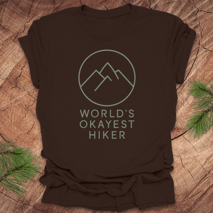 Brown t-shirt with 'World's Okayest Hiker' graphic on a wooden surface with pine branches.