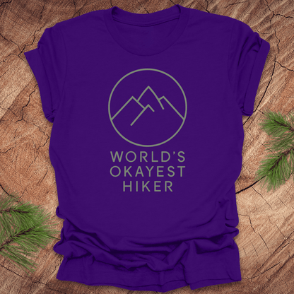 Purple t-shirt with mountain graphic and 'World's Okayest Hiker' text on a wooden surface.