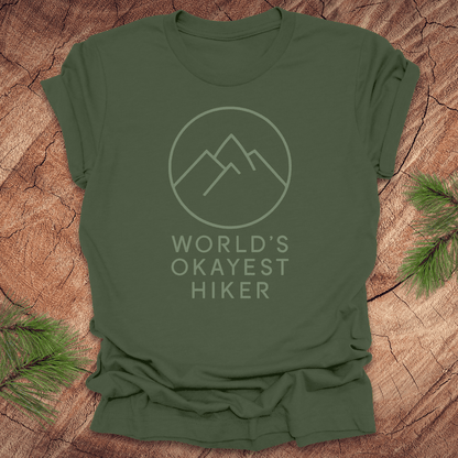 Green t-shirt with 'World's Okayest Hiker' text and mountain graphic on a wooden surface.