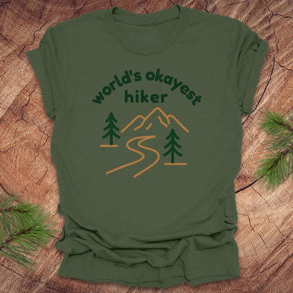 Green t-shirt with 'World's Okayest Hiker' text and mountain graphic on a wooden surface.