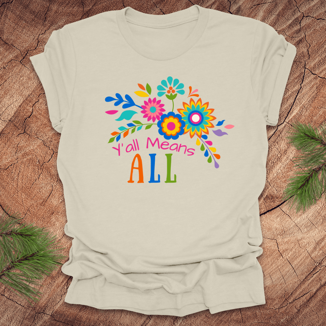 Beige t-shirt with colorful floral design and text 'Y'all Means All' on a wooden surface