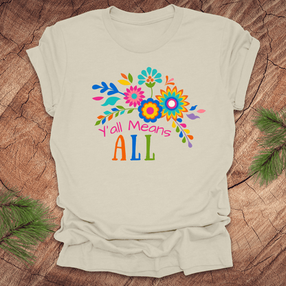 Beige t-shirt with colorful floral design and text 'Y'all Means All' on a wooden surface
