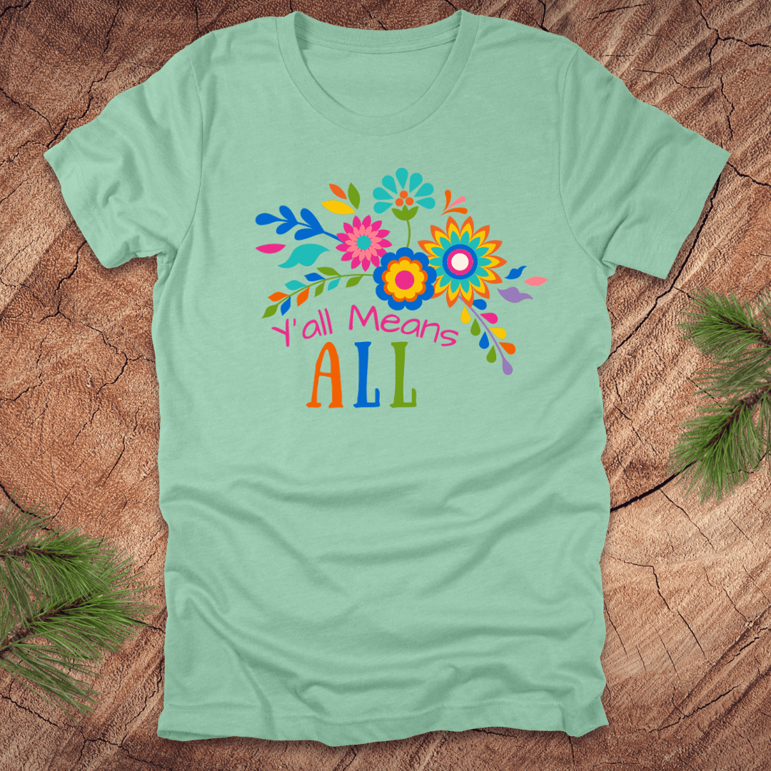 Mint green t-shirt with colorful floral design and text 'Y'all Means All' on a wooden surface