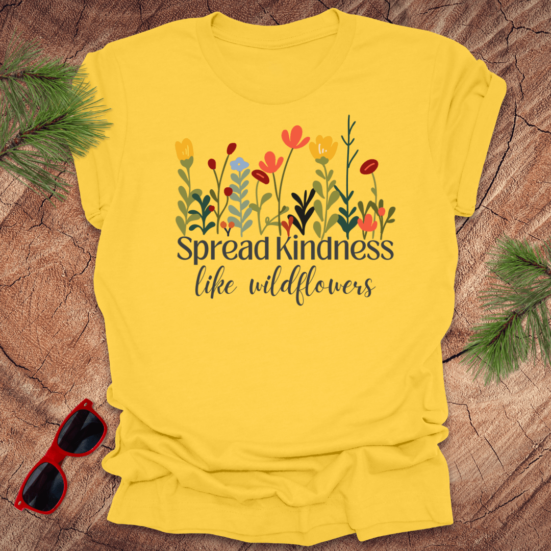 Yellow t-shirt with floral design and 'Spread kindness like wildflowers' text on a wooden surface with sunglasses and greenery.